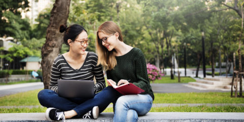 Two young female students, sitting on a brick wall outside, looking at a laptop computer.