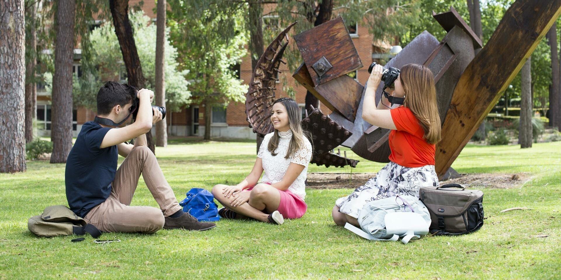 Humanities students holding a photoshoot, 2 students sitting on the grass with cameras while another models for them