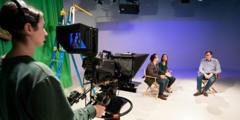 Three people sitting in a film studio, talking to each other, while a female camera operater is filming them