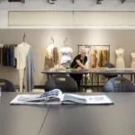 A blonde female fashion designer leaning over a desk, with a measuring tape in her hand
