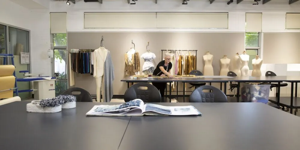 A blonde female fashion designer leaning over a desk, with a measuring tape in her hand