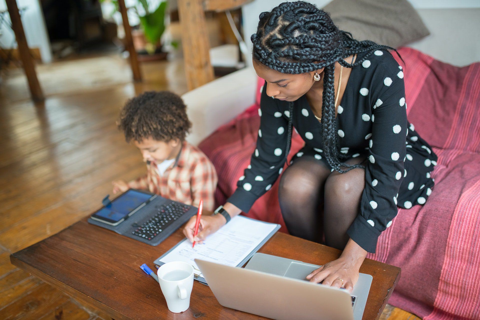 A person with long braided hair sits on a couch working on a laptop and writing in a notebook. A young child is sitting on the floor playing on a tablet beside them.