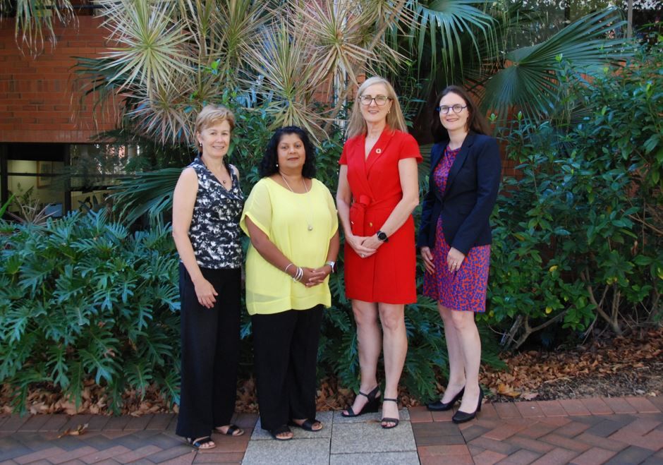 Four women stand in together in front of some bushes and a building