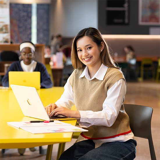 Smiling female student sitting at a yellow desk and looking up from her laptop