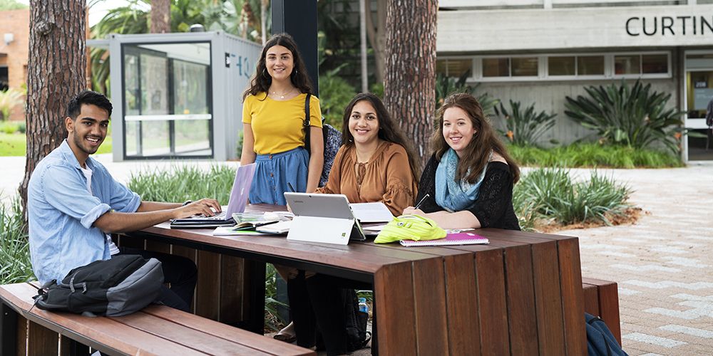 A group of students sitting on a park bench, looking at camera and smilig. 