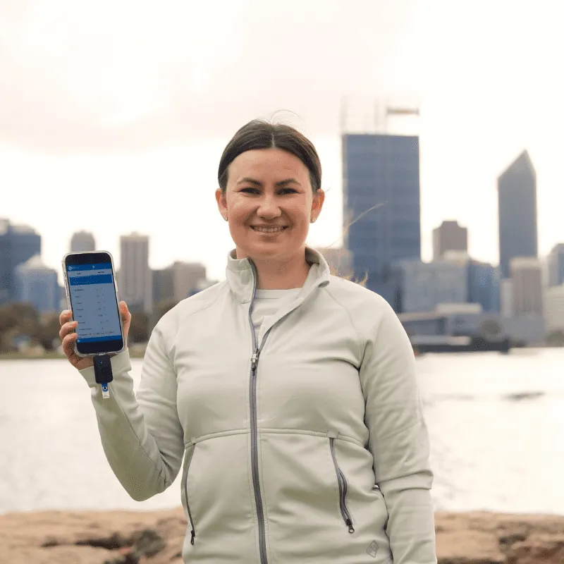 Person holding a smartphone near a waterfront with Perth city skyline in the background.