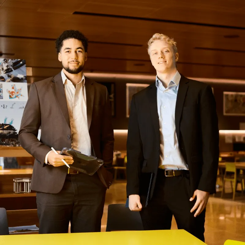 Two professionally dressed Indrus project team members standing in a modern office space, holding a tablet and folder, with tables and posters in the background.