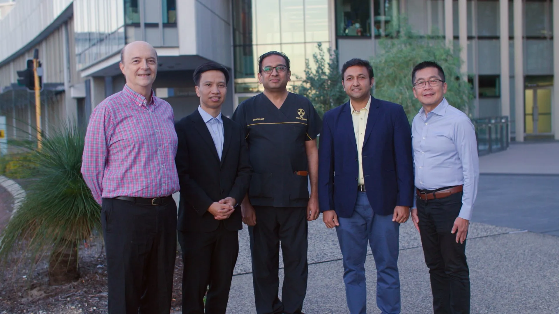 Five members of the Aura-CXR team stand in business and medical attire standing in front of a modern glass building.