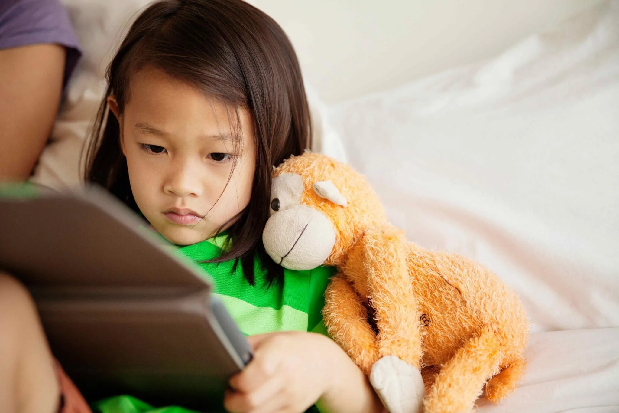 Child sitting on a bed or couch holding a tablet, with an orange stuffed monkey toy beside them.
