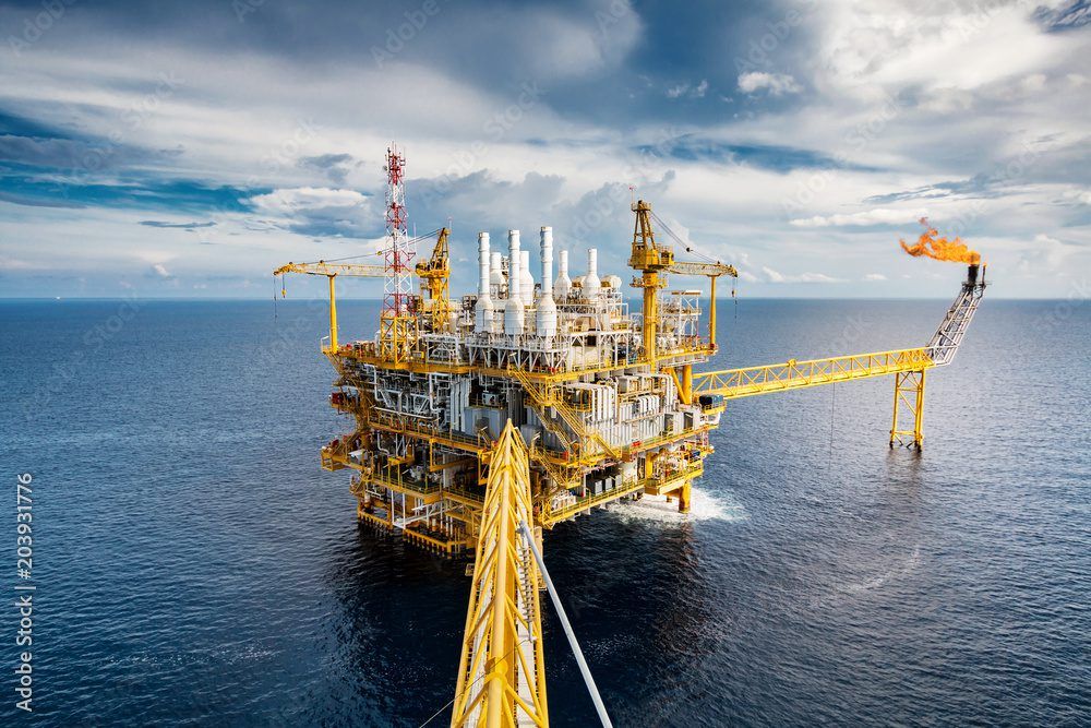 Offshore oil rig with cranes, pipelines, and a flare stack emitting flames, set against a partly cloudy sky and calm ocean.