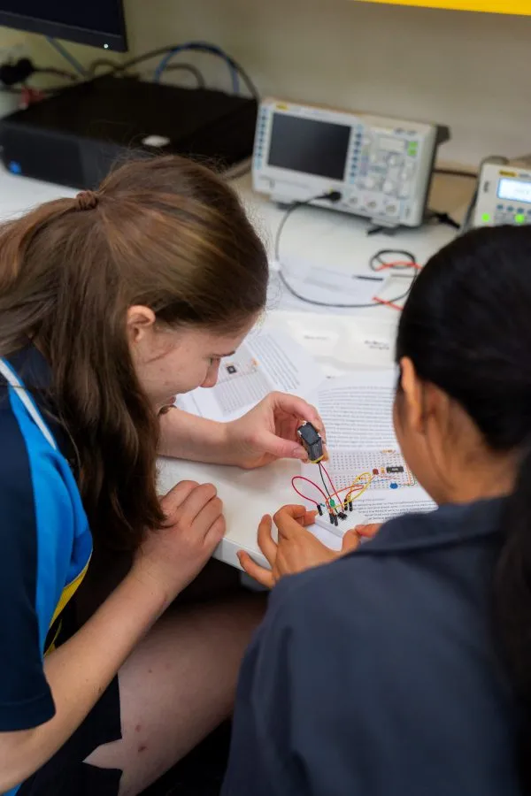 Two students work together at a classroom desk, assembling a small device with a battery and coloured wires. The desk behind them is covered with paper worksheets and other wires.