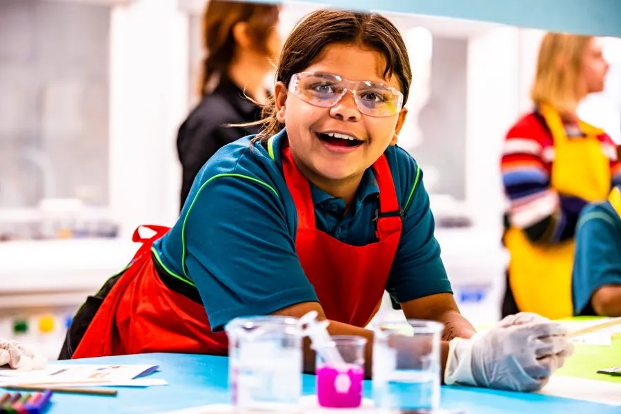 Girl wearing safety glasses and apron smiling at the camera while standing behind glass beakers in a lab.