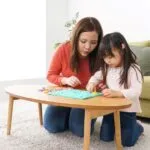 A woman and a young girl sit on the floor at a small round table, focusing together on a colorful board activity, suggesting a shared learning or playtime moment in a cozy home setting.