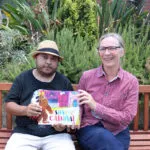 Man in a hat and a lady holding a book sitting on a bench.