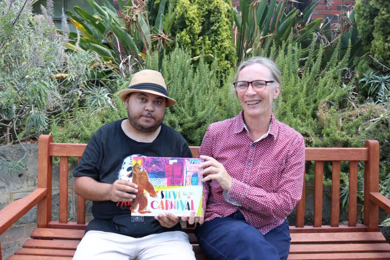 Man in a hat and a lady holding a book sitting on a bench.