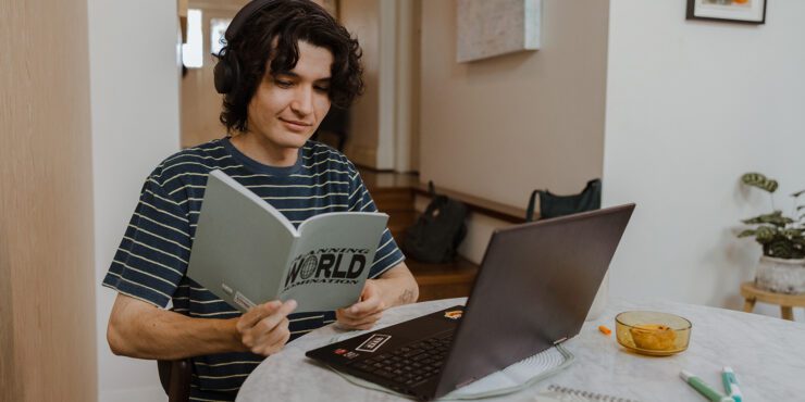 Student sitting at a round table reading a grey book with a black laptop in front of them.