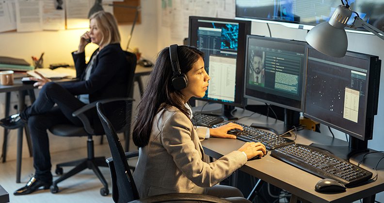 Serious female agent of intelligence service sitting in front of computer monitors in office and processing personal data of criminals