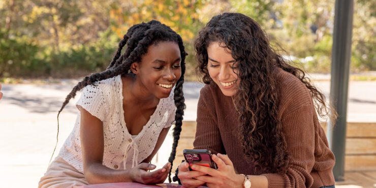 Two students sitting on a table, looking at the screen of one phone.