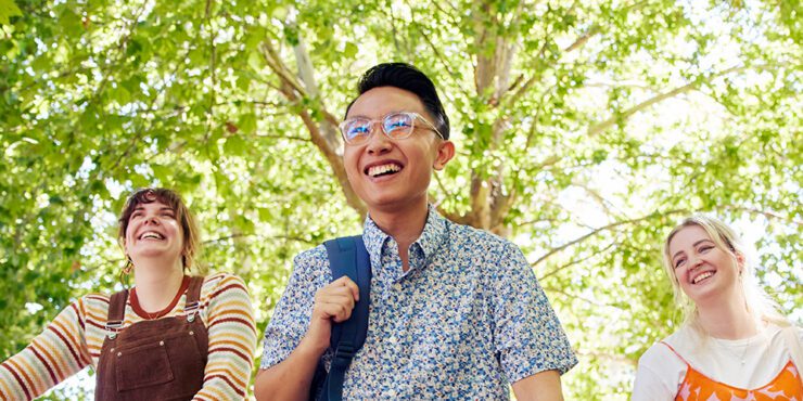 Three-students-walking in a line all smiling through the green trees on campus