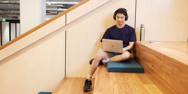 A male student sitting on some wooden steps looking at a laptop while wearing headphones