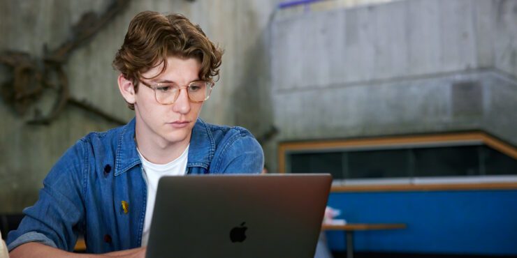 Student sitting indoors looking at a grey laptop.