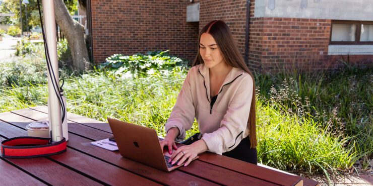 A female student sitting at an outdoor table and working on a laptop