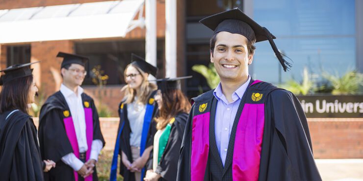 Young male graduate in graduation regalia and smiling and standing in front of a small group of graduates