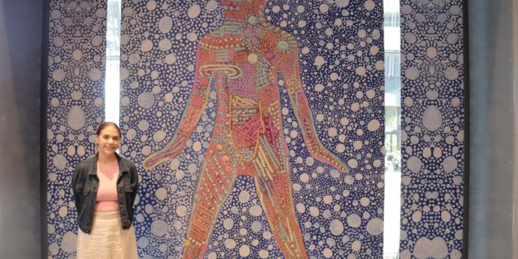 A student stands in front of the artwork she created, that features a red coloured figure on a blue patterned background.