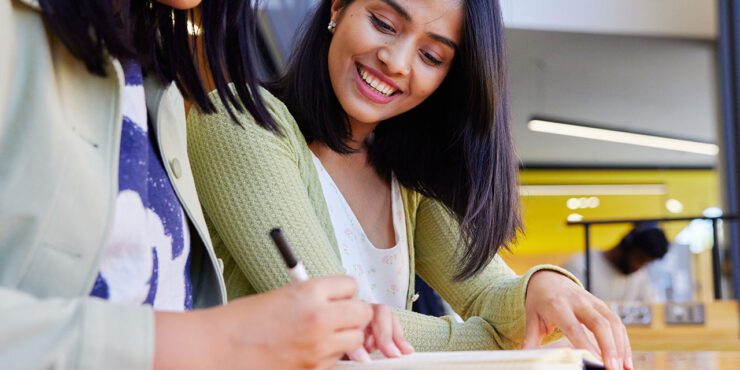 Two women looking down at notes, one smiling, one writing with a pen.