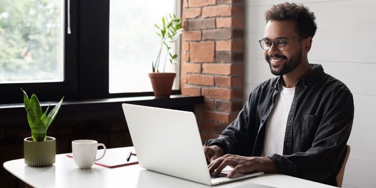 Young professional male, smiling, sitting at a desk at home and working on a laptop.