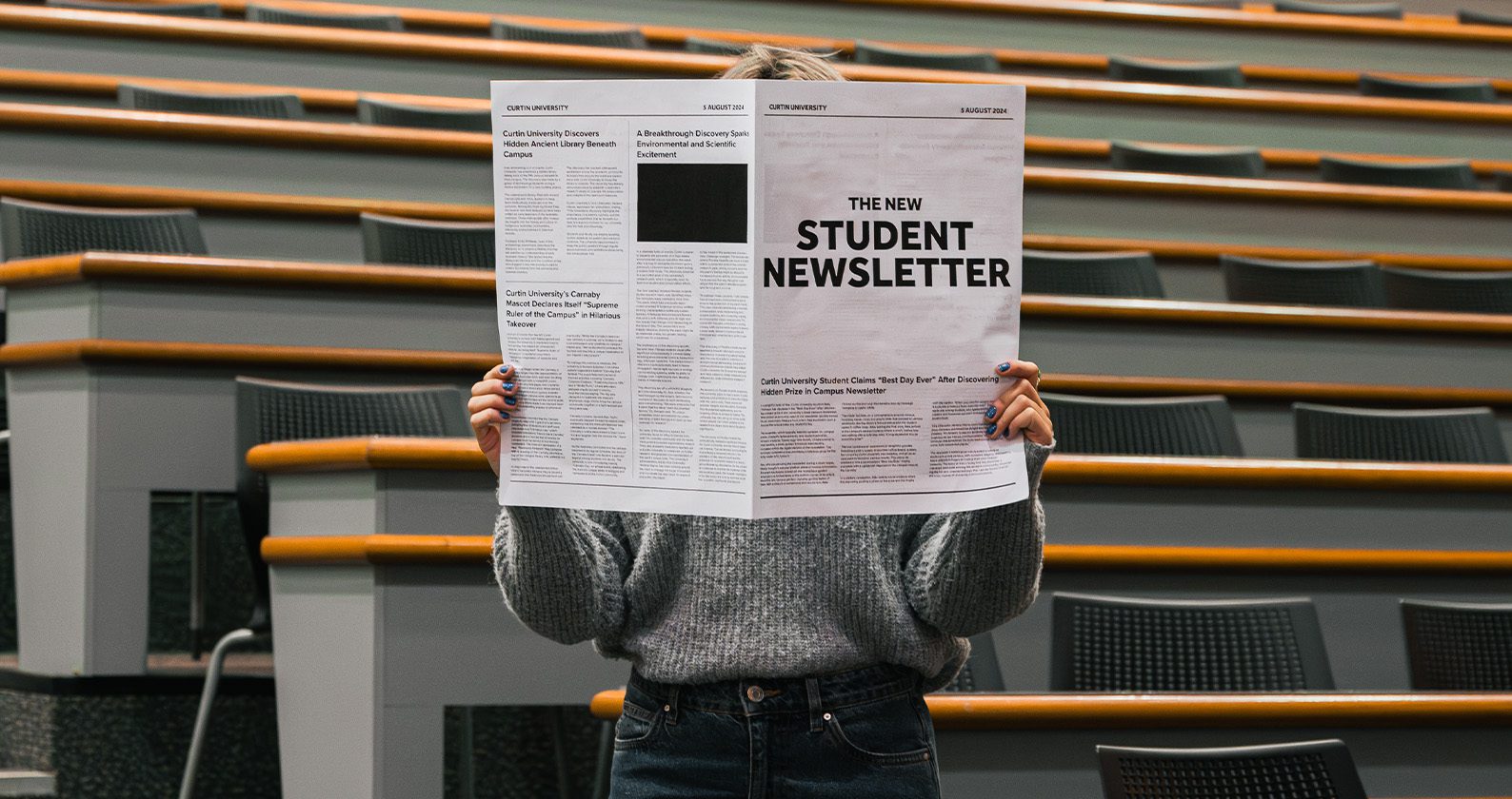 Person standing in a lecture theatre holding a newspaper in front of their face.