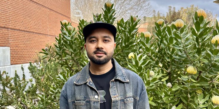 Success, a Master of Design student smiles for a photo wearing a denim jacket and black cap. Whilst standing in front of a pincushion bush.