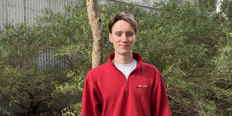 Pierce stands smiling at the camera, he wears a red jumper with a white t-shirt underneath. He is standing outside in front of foliage and trees.