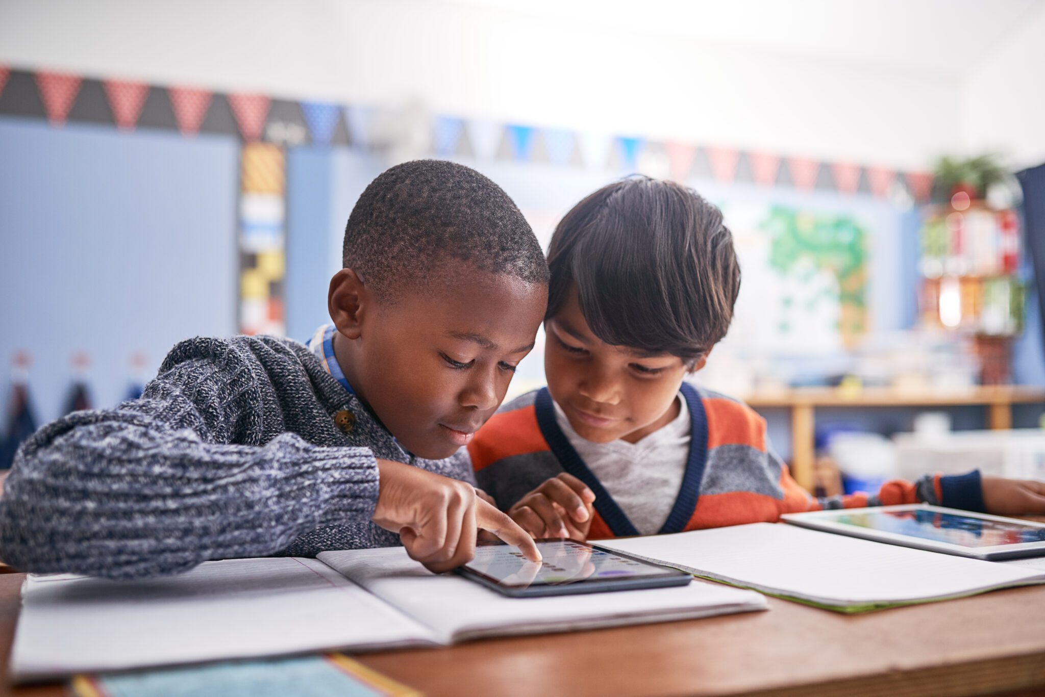 Two young boys with heads together look at a tablet in a classroom setting. 