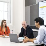 Three people seated around a table in a meeting room, discussing documents beside an open laptop, with a presentation slide displayed on a screen behind them.