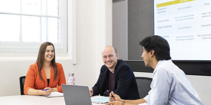 Three people seated around a table in a meeting room, discussing documents beside an open laptop, with a presentation slide displayed on a screen behind them.
