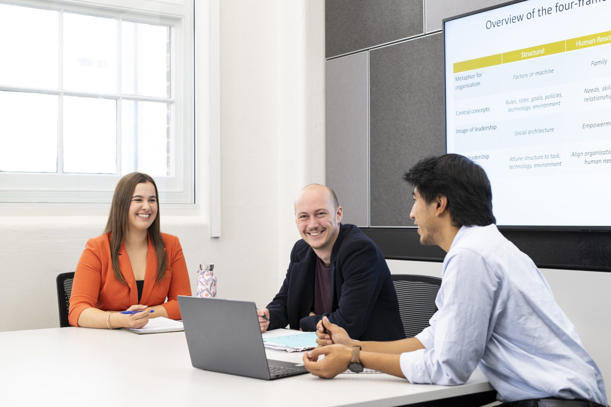 Three people seated around a table in a meeting room, discussing documents beside an open laptop, with a presentation slide displayed on a screen behind them.