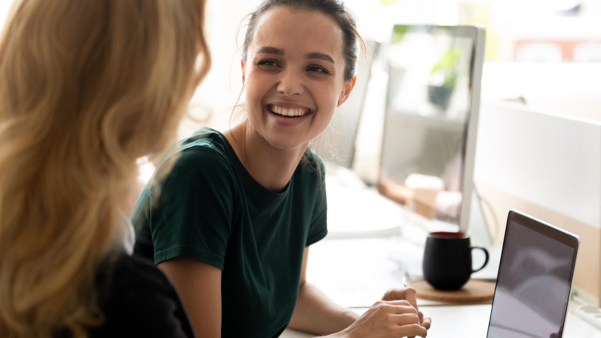 Two people sitting at a desk in an office setting, engaged in conversation. A laptop is open on the desk, and a black coffee mug is placed on a wooden coaster beside it. A large computer monitor is visible in the background.