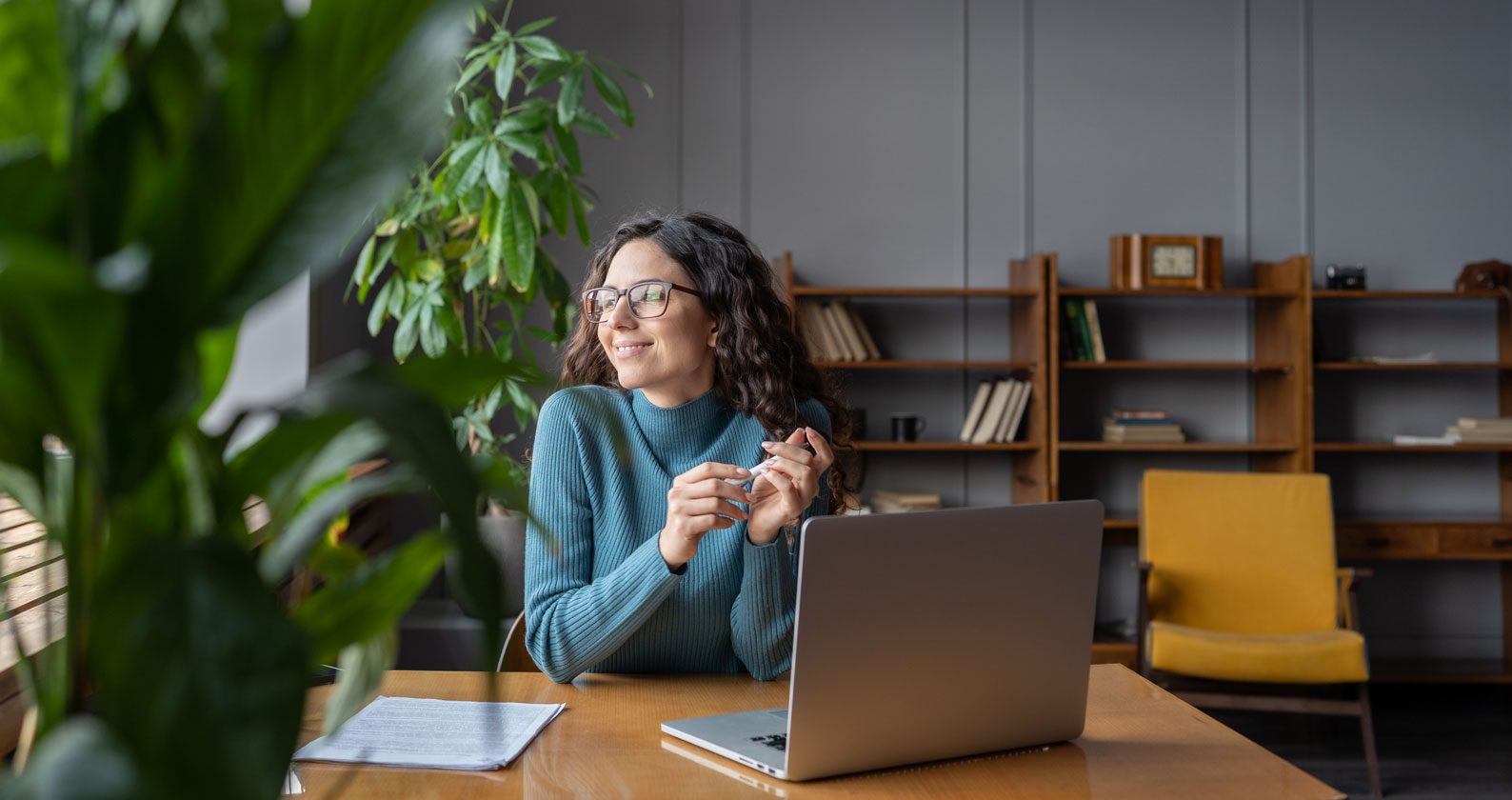 A woman looking off into the distance while sitting at a table with a laptop.