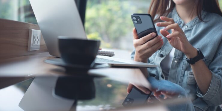 On a bench, there is a touch-screen device, a coffee and a laptop. A person sits at the bench, in the background, on their phone.