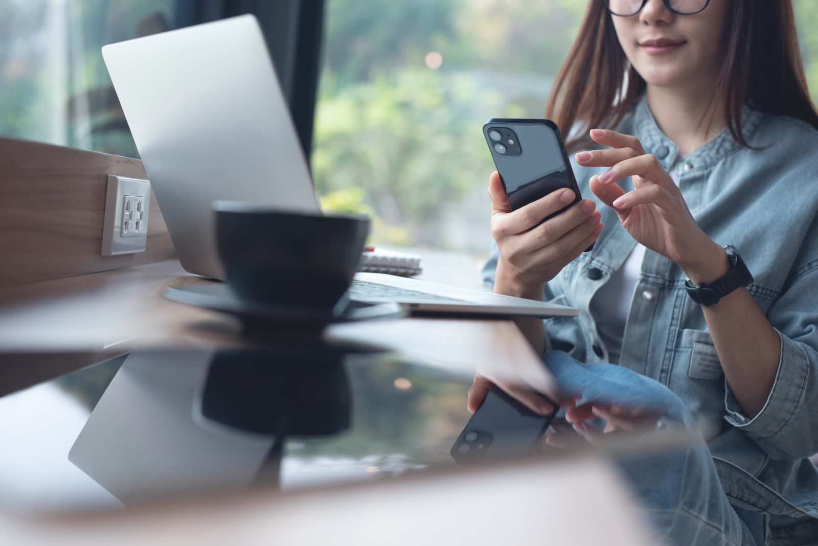 On a bench, there is a touch-screen device, a coffee and a laptop. A person sits at the bench, in the background, on their phone.