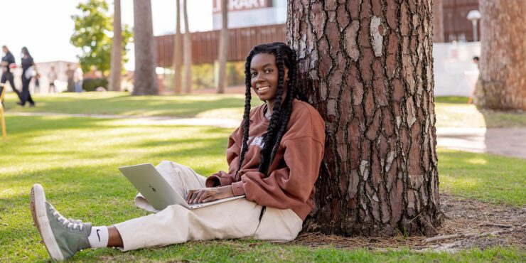 A student sitting on the grass, leaning on a tree with her laptop open.