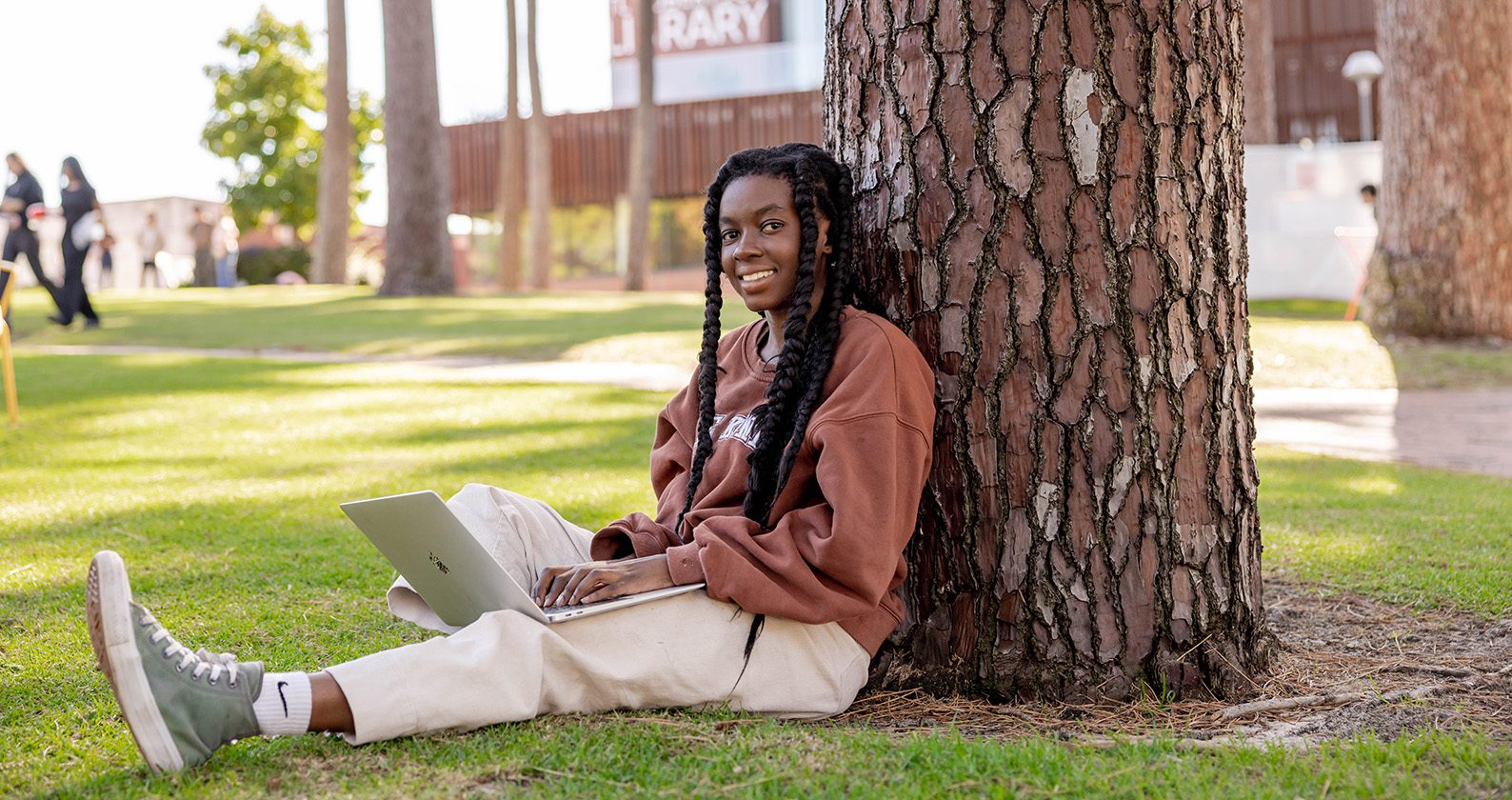 A student sitting on the grass, leaning on a tree with her laptop open.