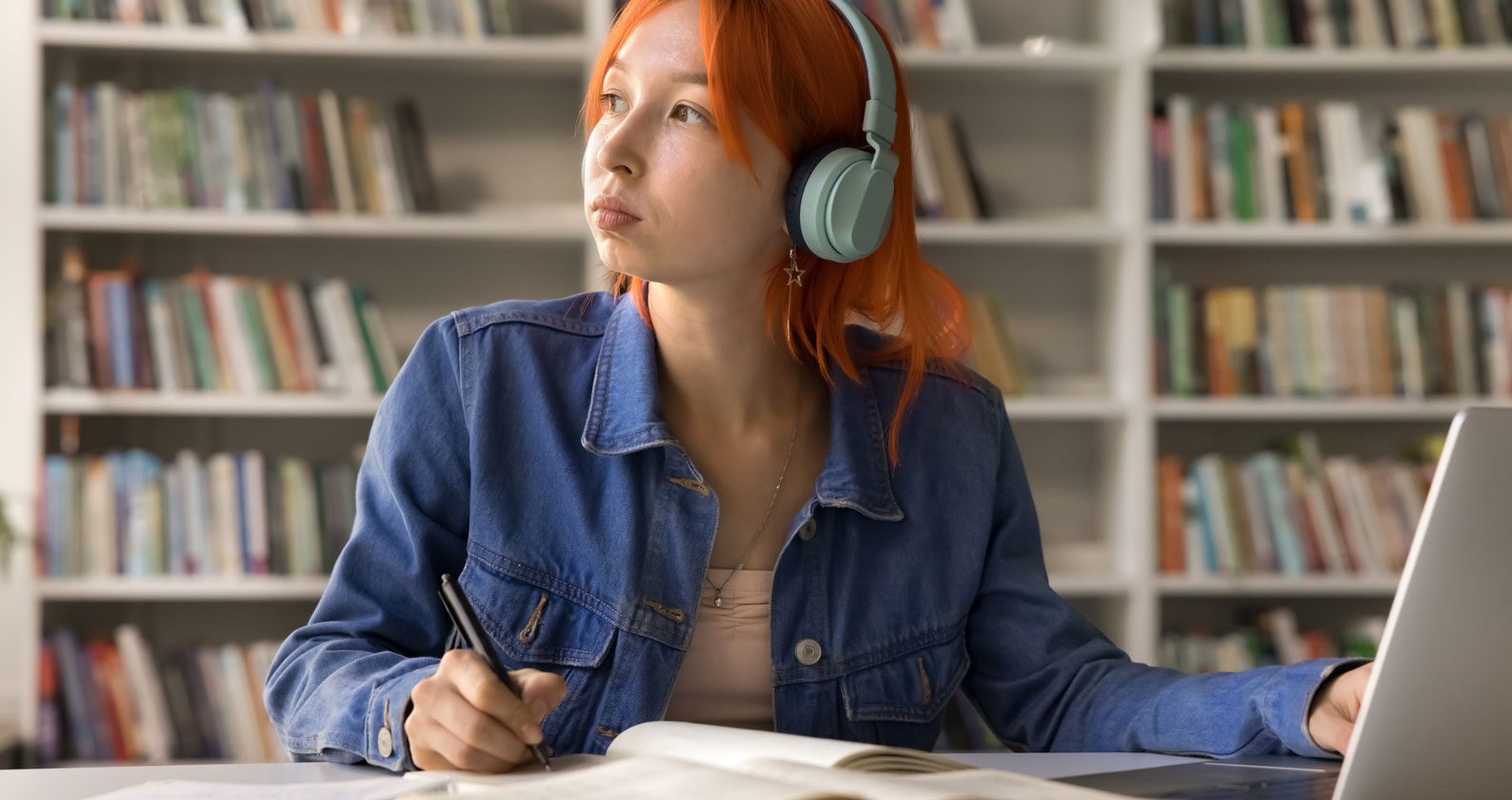 Student looking off into the distance wearing headphones while sitting at a desk with a laptop.