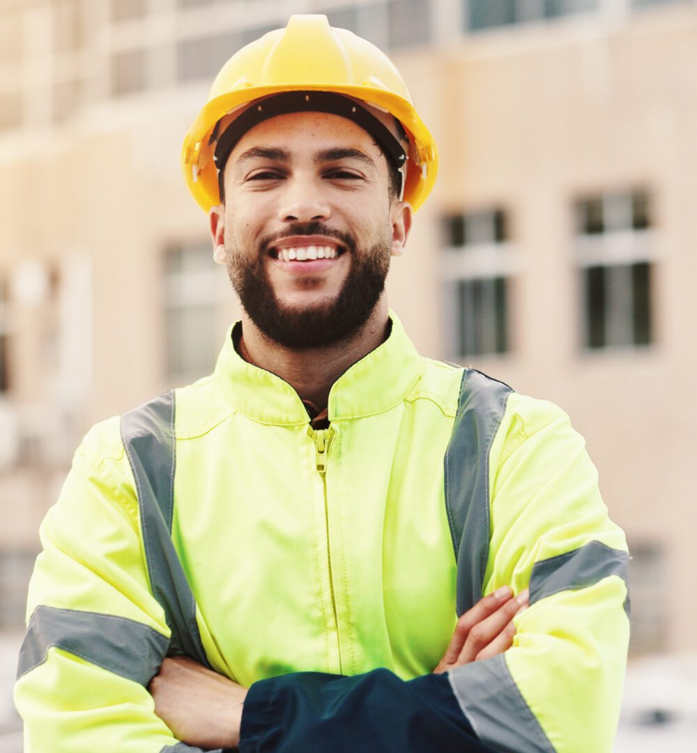 Man with a hard hat in foreground of a construction site, arms crossed and smiling.