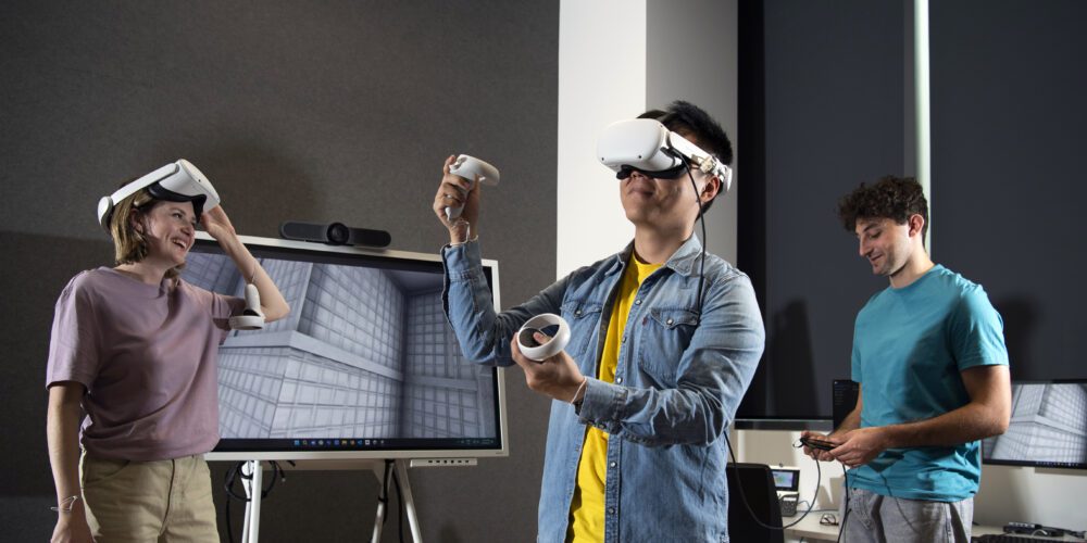 Three students explore virtual reality in a modern classroom. One wears a VR headset while others interact with controllers and digital displays.
