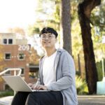 A smiling student sits outdoors on campus with a laptop, surrounded by trees and a building marked “209” in the background.