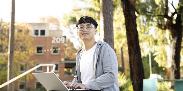 A smiling student sits outdoors on campus with a laptop, surrounded by trees and a building marked “209” in the background.