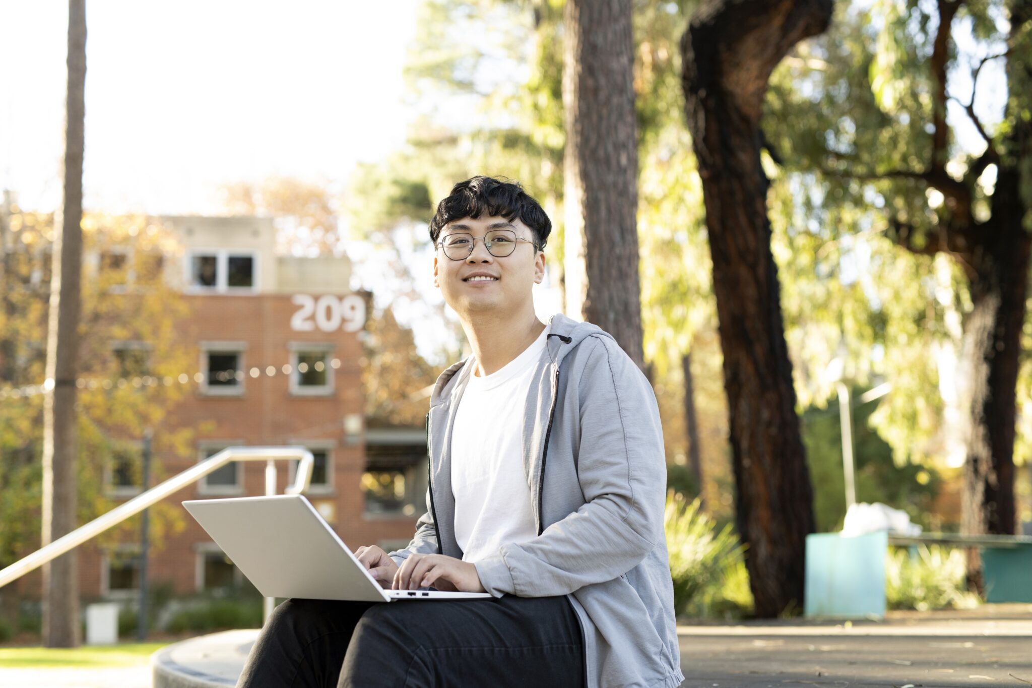 A smiling student sits outdoors on campus with a laptop, surrounded by trees and a building marked “209” in the background.