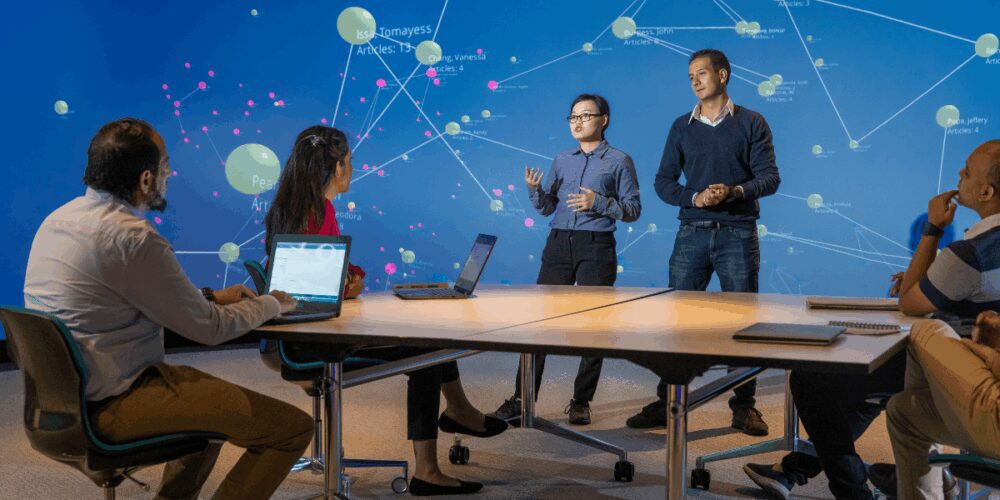 Two students presenting in front of a large interactive data visualisation screen, while classmates watch from their desks.